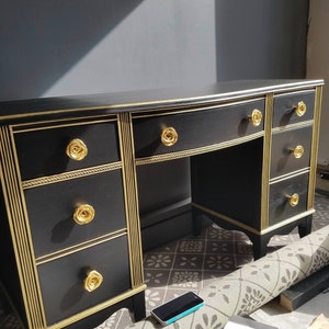 以下が含まれることがあります： 金色の縁取りとバラの形をした引き出しの取っ手がついた黒い机。机は前面が湾曲していて、引き出しが複数あります。机は模様のある敷物の上に置かれています。