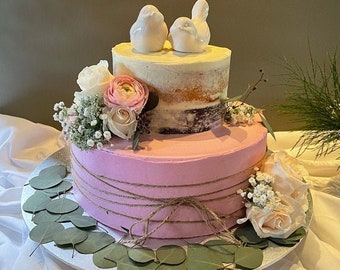 May include: A two-tiered wedding cake with a pink bottom layer and a white top layer. The cake is decorated with a rustic twine bow, fresh flowers, and two white ceramic birds on top.