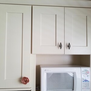 以下が含まれることがあります： 黒いダイヤモンド型のノブが付いた白いキッチンのキャビネット。キャビネットの下には白い電子レンジがあります。左側のキャビネットのドアには、ローズゴールドのノブが付いています。