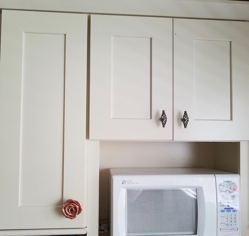 以下が含まれることがあります： 黒いダイヤモンド型のノブが付いた白いキッチンのキャビネット。キャビネットの下には白い電子レンジがあります。左側のキャビネットのドアには、ローズゴールドのノブが付いています。