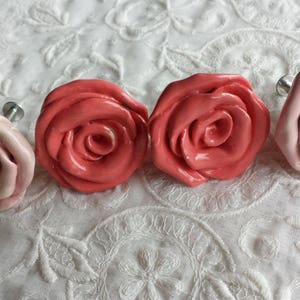 K&ouml;nnte beinhalten: Vier Keramik-Schubladengriffe in Rosenform in den Farben Rosa und Koralle. Die Griffe haben eine silberne Metallschraubenbefestigung.