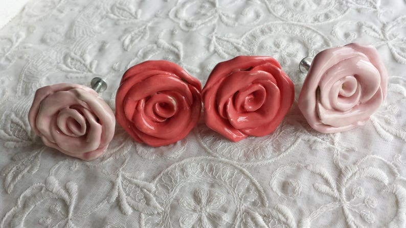 K&ouml;nnte beinhalten: Vier Keramik-Schubladengriffe in Rosenform in den Farben Rosa und Koralle. Die Griffe haben eine silberne Metallschraubenbefestigung.