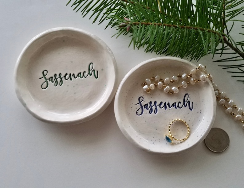 May include: Two white ceramic trinket dishes with the word "Sassenach" written in green and blue lettering. One dish has a pearl necklace and a ring with a blue stone on it.