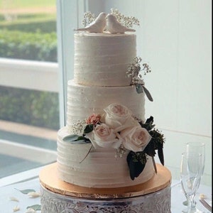 May include: A three-tiered white frosted cake with horizontal textured icing. The top tier features two white bird figurines and baby's breath. The middle tier is adorned with light pink roses, greenery, and baby's breath on a gold cake board.