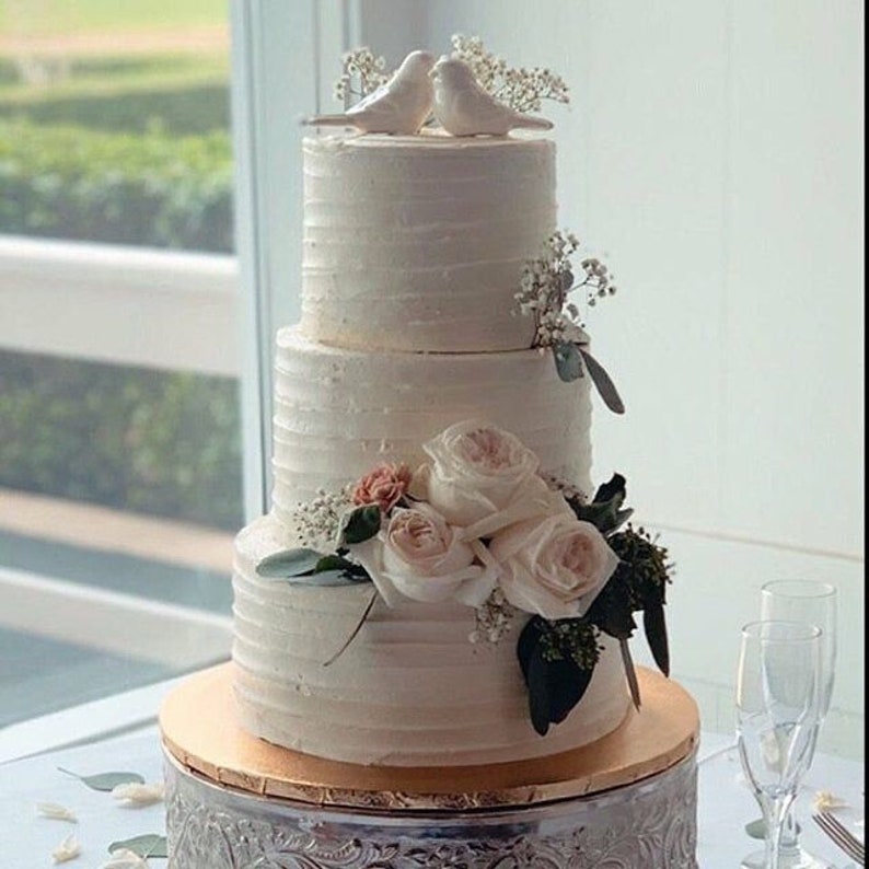 May include: A three-tiered white frosted cake with horizontal textured icing. The top tier features two white bird figurines and baby's breath. The middle tier is adorned with light pink roses, greenery, and baby's breath on a gold cake board.