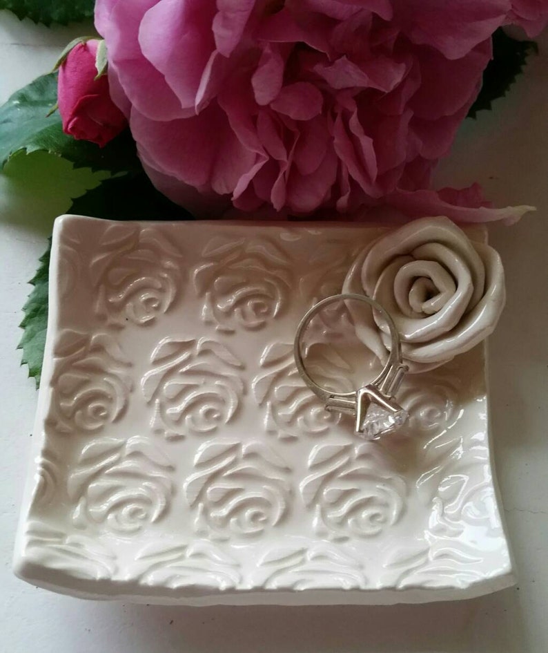 May include: White ceramic ring dish with a raised rose pattern. The dish is shaped like a rectangle with rounded corners. A silver ring with a large diamond is sitting on the dish.