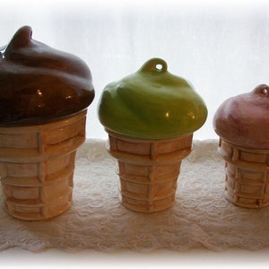 May include: Three ceramic ice cream cone shaped containers with lids. The lids are different colors: brown, green, and pink. The cones are all the same size and have a waffle pattern.