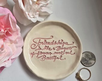 May include: A white ceramic trinket dish with a pink inscription that reads "Friendship is like a Bouquet of flowers, sweet and beautiful."