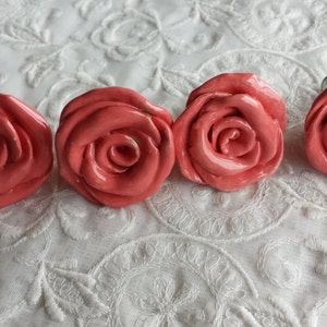 May include: Four coral pink ceramic rose-shaped drawer pulls. The pulls have a glossy finish and are arranged in a row.