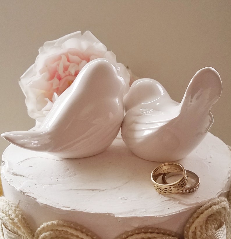 May include: Two white ceramic birds perched on a white cake with a white rose and two gold wedding bands.