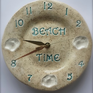 May include: A beige ceramic clock with a speckled finish. The clock face features the words "Beach Time" and seashell accents. The clock hands are gold.