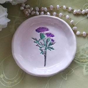 May include: A white ceramic trinket dish with a raised design of a purple thistle flower.