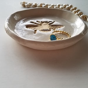 May include: White ceramic ring dish with a gold bee design and a blue ring inside.