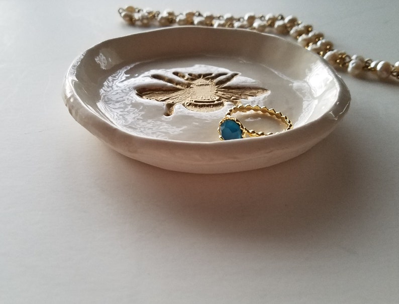 May include: White ceramic ring dish with a gold bee design and a blue ring inside.