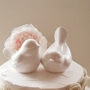 May include: White ceramic bird cake topper set. The birds are facing each other and are perched on a white cake with a beige lace trim.