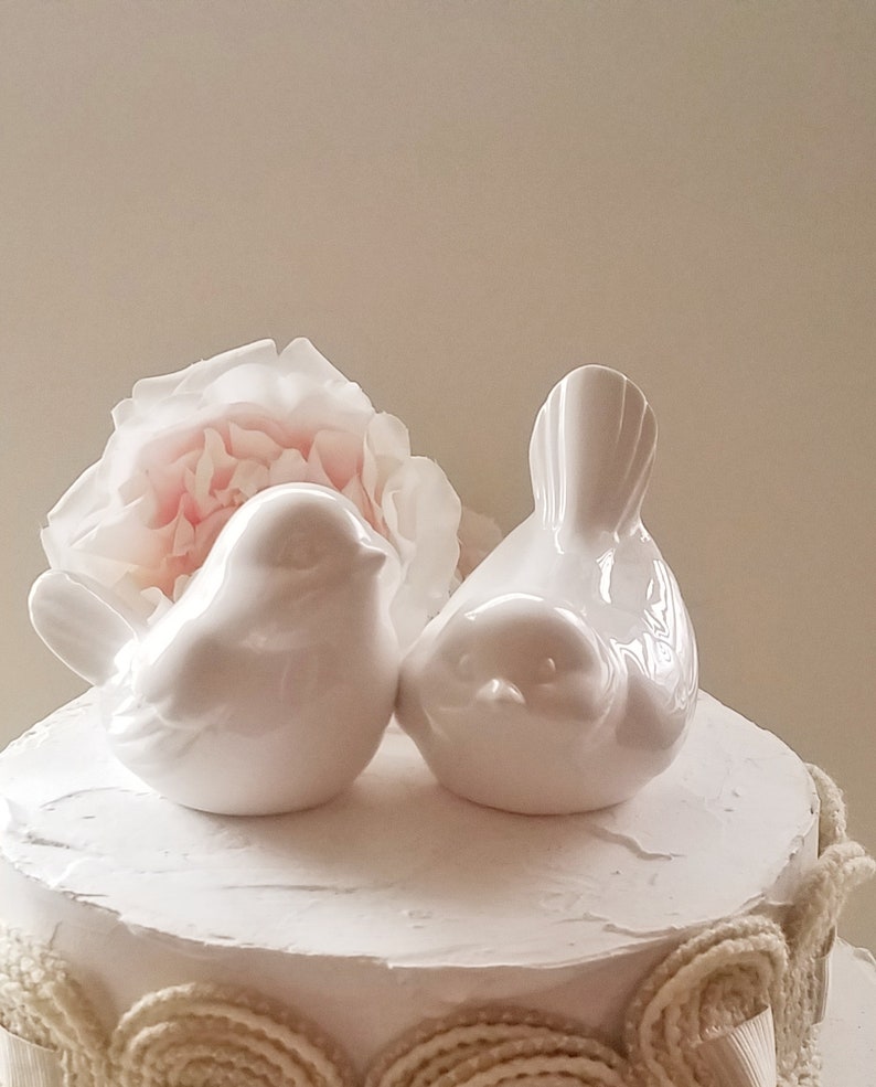 May include: White ceramic bird cake topper set. The birds are facing each other and are perched on a white cake with a beige lace trim.