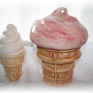 May include: Two ceramic ice cream cone shaped containers. The larger cone is pink and white with a swirl design. The smaller cone is white with a brown waffle cone.