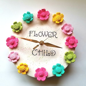 May include: A white ceramic clock with a gold clock hand and the words "FLOWER CHILD" in black lettering. The clock face is decorated with colorful flower-shaped accents.