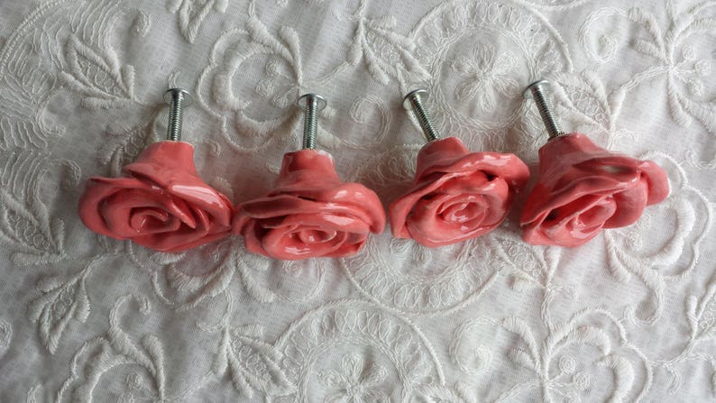 K&ouml;nnte beinhalten: Vier rosafarbene Keramik-Schubladengriffe in Rosenform mit silberner Metallbeschl&auml;ge.