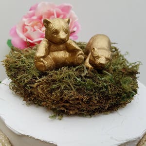 Puede incluir: Dos figuritas de oso pintadas de oro sentadas en un lecho de musgo verde. Los osos están en una pose relajada, uno sentado erguido y el otro acostado. El musgo está rodeado de un pastel blanco.