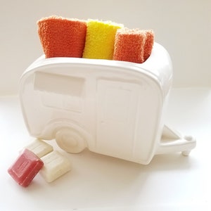 May include: A white ceramic camper-shaped holder containing three folded terry cloth sponges in orange, yellow, and orange. Three small square soap bars in red and white are positioned on the surface in front of the camper-shaped holder.