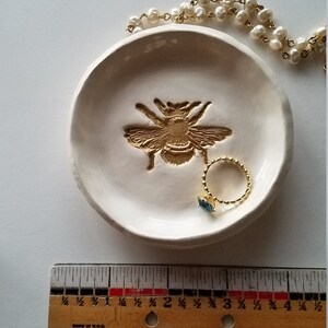 May include: White ceramic ring dish with a gold bee design. A gold ring with a blue stone is sitting in the dish.