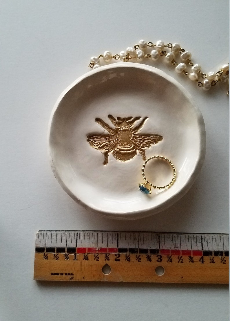 May include: White ceramic ring dish with a gold bee design. A gold ring with a blue stone is sitting in the dish.