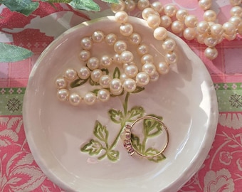 May include: A cream-colored ceramic ring dish with a floral design and a gold ring with pink stones. A strand of faux pearls rests in the dish, and another is draped nearby. The background features a pink floral pattern.