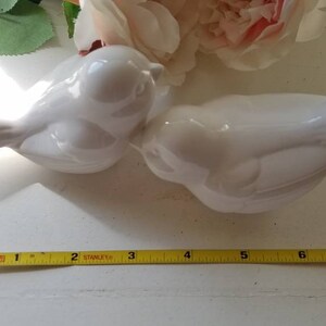May include: Two white ceramic bird figurines, one facing left and the other facing right, are positioned on a white surface. The birds are kissing, with their beaks touching. A yellow measuring tape is visible at the bottom of the image.