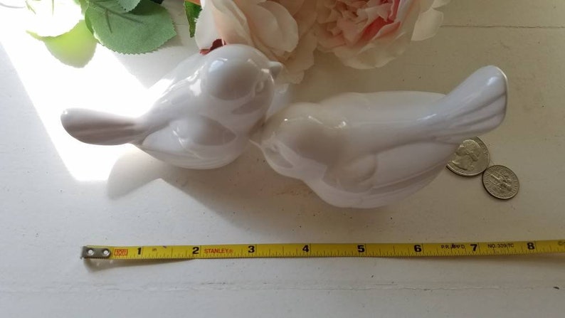 May include: Two white ceramic bird figurines, one facing left and the other facing right, are positioned on a white surface. The birds are kissing, with their beaks touching. A yellow measuring tape is visible at the bottom of the image.