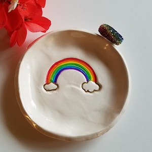 May include: White ceramic ring dish with a rainbow design and a rainbow-colored ring on the side.