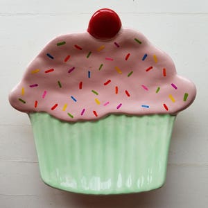 May include: A ceramic cupcake-shaped dish featuring a light green fluted base and a pink frosting top decorated with multi-colored sprinkles. A glossy red cherry sits at the very center of the pink frosting, completing the dessert-themed design.