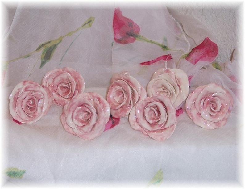 May include: A set of eight ceramic rose-shaped drawer pulls in a pale pink color. The pulls are arranged on a white fabric background.