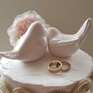 May include: Two white ceramic birds perched on a white cake with a pink flower. Two gold wedding bands are placed on the cake.