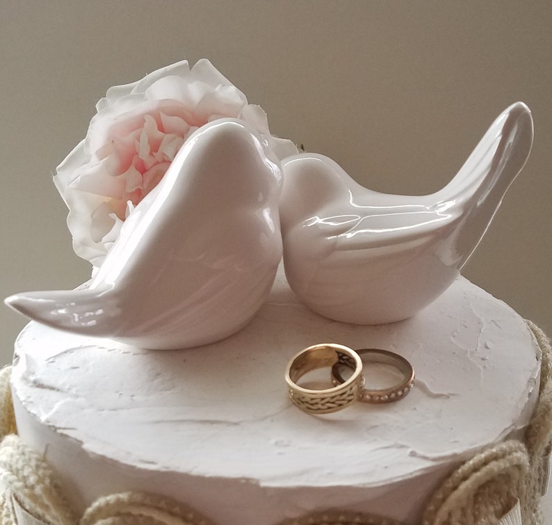 May include: Two white ceramic birds perched on a white cake with a pink flower. Two gold wedding bands are placed on the cake.
