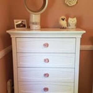 May include: White dresser with four drawers and pink ceramic knobs. The dresser has a slightly distressed finish and is made of wood.