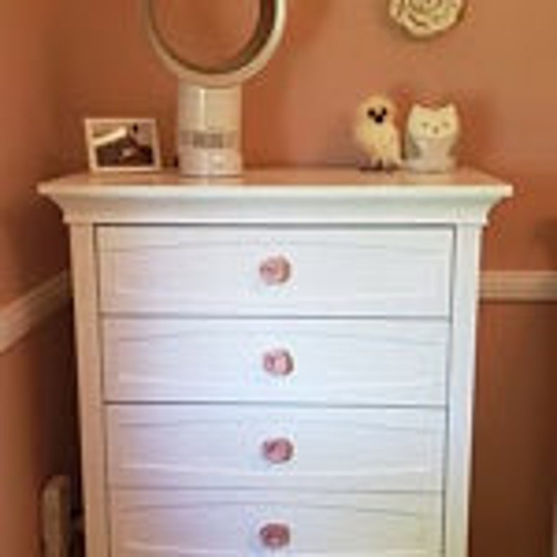 May include: White dresser with four drawers and pink ceramic knobs. The dresser has a slightly distressed finish and is made of wood.