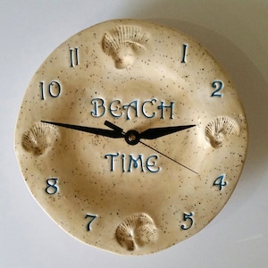 May include: A round ceramic wall clock with a speckled cream finish, featuring blue numerals from 1 to 12 and the text "BEACH TIME" in the center. The clock face is decorated with four embossed seashell motifs and has black metal clock hands.