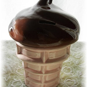 May include: A ceramic ice cream cone piggy bank with a brown chocolate-colored top and a pink and white waffle cone base.