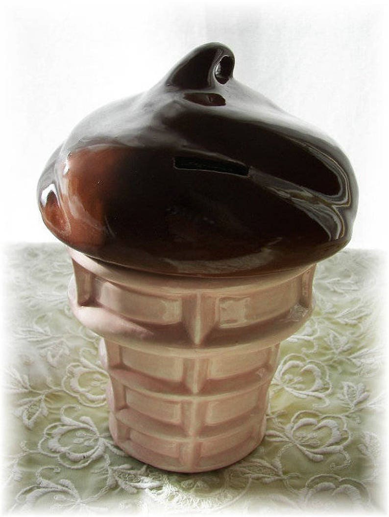 May include: A ceramic ice cream cone piggy bank with a brown chocolate-colored top and a pink and white waffle cone base.