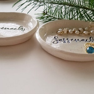 May include: Two white ceramic trinket dishes with the word "Jazzercise" written on them in black script. One dish has a pearl necklace and a gold ring with a blue stone.