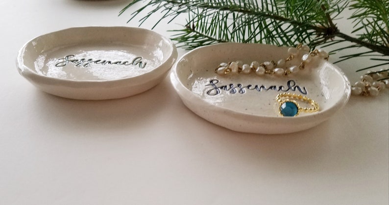 May include: Two white ceramic trinket dishes with the word "Jazzercise" written on them in black script. One dish has a pearl necklace and a gold ring with a blue stone.