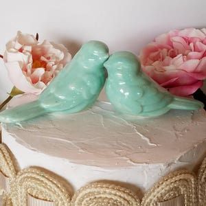 May include: A pair of light blue ceramic birds perched on top of a white cake with a pink and white frosting design. The birds are facing each other with their beaks touching.