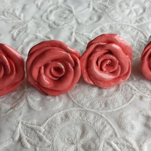 May include: Four coral pink ceramic rose-shaped drawer pulls. The pulls have a glossy finish and are arranged in a row on a white background.