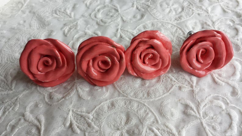 K&ouml;nnte beinhalten: Vier korallenrosa Keramik-Rosenkn&ouml;pfe. Die Kn&ouml;pfe haben eine gl&auml;nzende Oberfl&auml;che und sind in einer Reihe auf einem wei&szlig;en Hintergrund angeordnet.