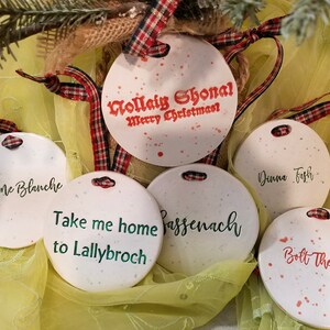 May include: Five white round ceramic ornaments with red and green text. The ornaments feature phrases from the popular TV show "Outlander". The phrases include "La Dame Blanche", "Nollaig Shonal Merry Christmas", "Take me home to Lallybroch", "Bassenach", and "Bolt the Door".
