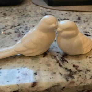 May include: Two glossy white ceramic bird figurines positioned side by side. The left bird is elongated with a long tail, while the right bird is rounder and more compact. Both birds have smooth, featureless surfaces and are joined together.