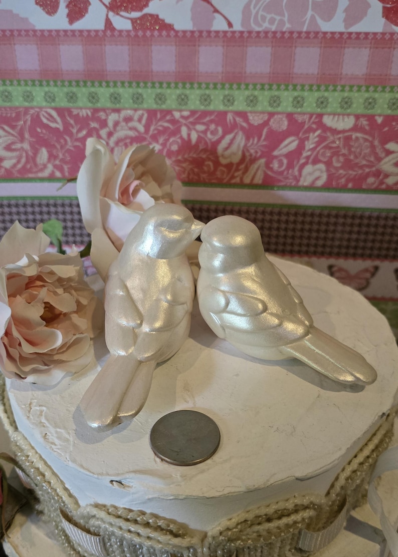 May include: Two pearlescent white bird figurines positioned side-by-side on a circular white base. A round silver coin is placed between the birds for scale. The birds feature smooth, glossy surfaces and detailed wing carvings.