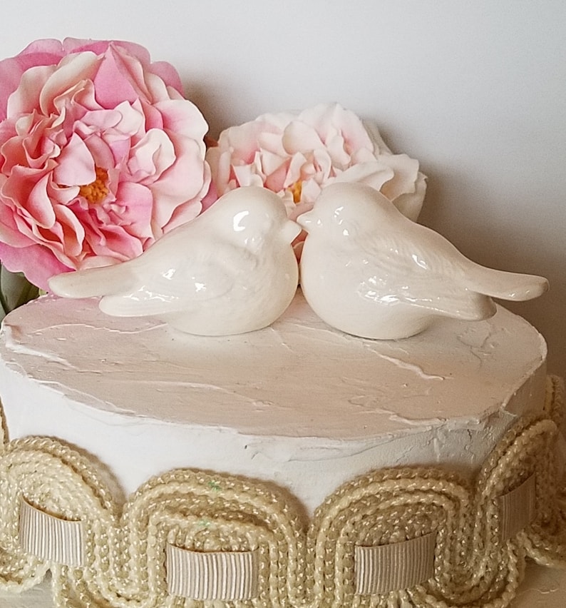 May include: White ceramic bird cake topper with two birds facing each other. The birds are perched on a white cake with a beige lace trim.
