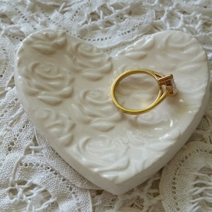 May include: A heart-shaped, off-white ceramic ring dish with a raised rose pattern. A gold ring with a square, clear gemstone rests in the center of the dish. The dish is on a white lace surface.